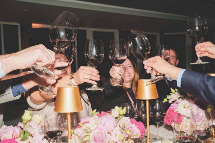 Guests raise glasses of red wine during an Artisans of Wine & Food dinner at The Boca Raton, surrounded by pink flowers and gold table lamps.