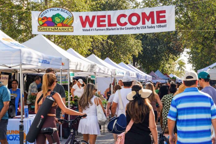 Shoppers beneath the West Palm Beach GreenMarket welcome banner, with text announcing its October 3 return.