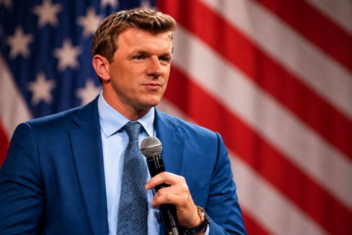 James O'Keefe holding a microphone in front of an American flag ahead of his October 27 Palm Beach appearance